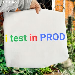 May include: A white tote bag with the words "i test in PROD" printed in colorful letters. The text is in yellow, green, red, and blue. The bag is held up by a person's hand.