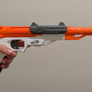 May include: A Nerf Sharpfire toy blaster featuring a bright orange barrel and front sight, a white handle and frame, and a dark gray top-mounted tactical rail. The Nerf logo is printed in black on the side of the white frame, and the word Sharpfire is embossed on the gray rail component.