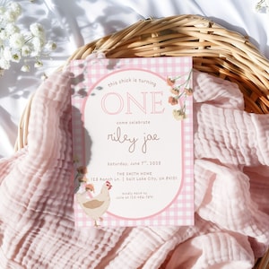 Puede incluir: Una tarjeta de invitación rosa y blanca con el texto "this chick is turning ONE" y "riley jae" en cursiva. La tarjeta tiene un patrón de cuadros y una ilustración de un pollo de dibujos animados. La invitación está colocada en una cesta de mimbre con tela rosa.