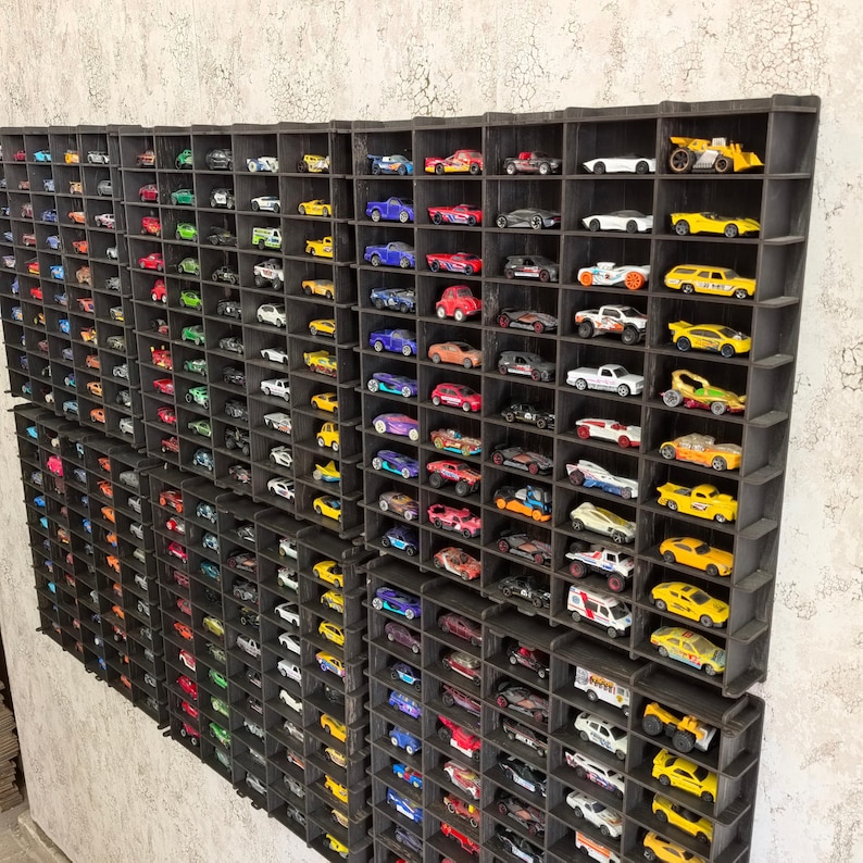 Op de afbeelding: Een wandrek met meerdere vakken, elk met een klein speelgoedautootje. De auto's zijn gerangschikt in rijen en kolommen, en ze zijn allemaal in verschillende kleuren en stijlen. Het rek is gemaakt van zwart hout en heeft een rustieke uitstraling.