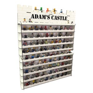 Puede incluir: Una estantería de madera blanca con un diseño de pared de ladrillo y el texto "ADAM'S CASTLE" en la parte superior. La estantería tiene varios estantes de acrílico transparente para exhibir minifiguras de Lego.