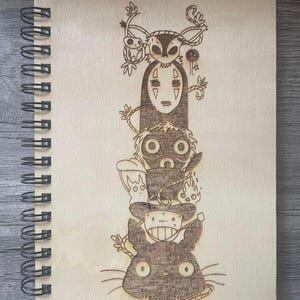 Puede incluir: Un cuaderno de espiral de madera con un dise&ntilde;o grabado con l&aacute;ser de personajes de la pel&iacute;cula de animaci&oacute;n El viaje de Chihiro. El dise&ntilde;o presenta a Sin Cara, el gato negro y otros personajes de la pel&iacute;cula.