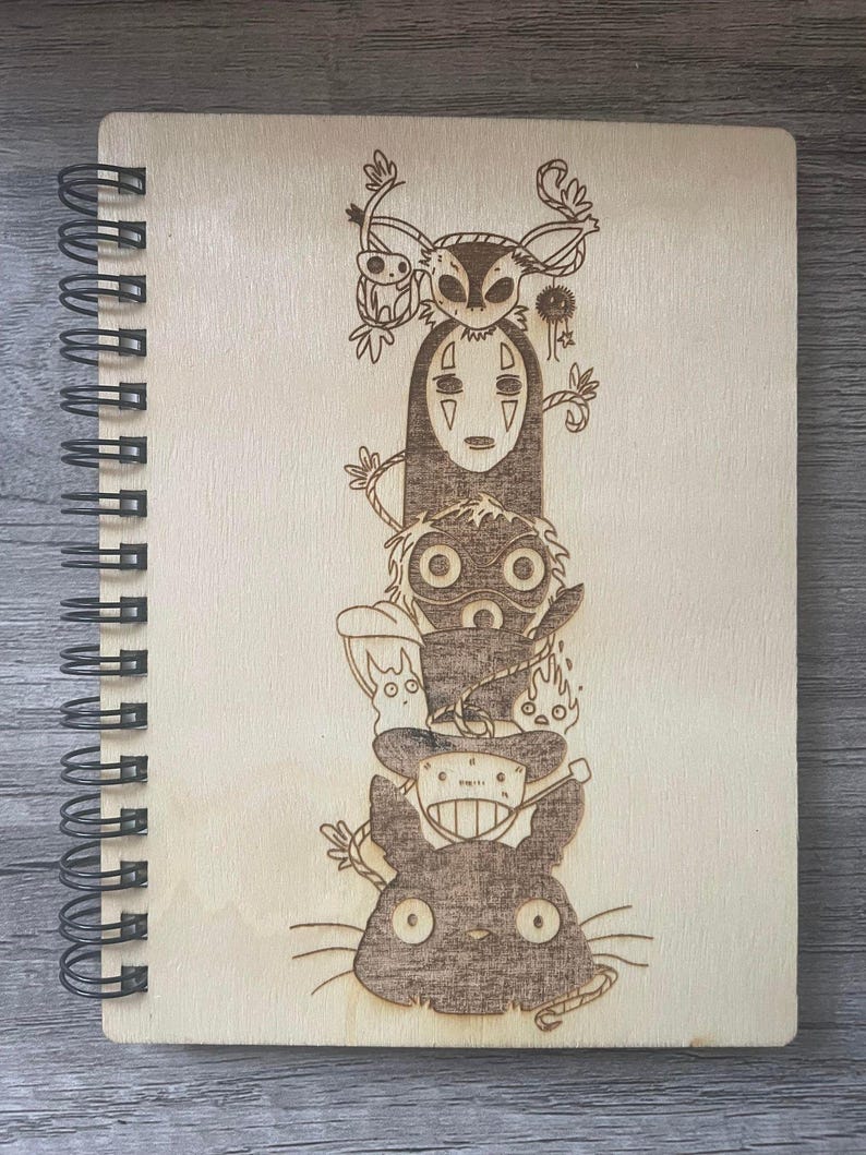 Puede incluir: Un cuaderno de espiral de madera con un dise&ntilde;o grabado con l&aacute;ser de personajes de la pel&iacute;cula de animaci&oacute;n El viaje de Chihiro. El dise&ntilde;o presenta a Sin Cara, el gato negro y otros personajes de la pel&iacute;cula.