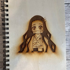 May include: A wooden spiral-bound notebook with a laser-etched image of an anime character. The character has long dark hair, large eyes, and a bamboo muzzle. The notebook's cover is light-colored wood, and the image is a darker brown.