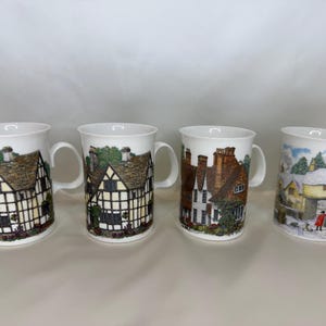 May include: Four white ceramic mugs, each featuring a different detailed village scene. The mugs have a curved handle and a white interior. The village scenes depict houses, trees, and people. The mugs are displayed on a white surface.