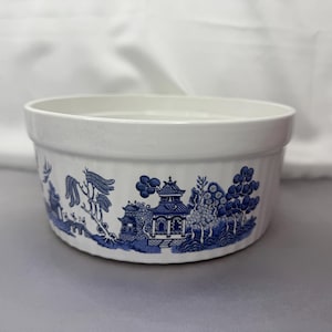 Op de afbeelding: Een witte keramische bakvorm met een blauw en wit wilgenmotief. Het patroon toont een pagode, bomen en een brug.