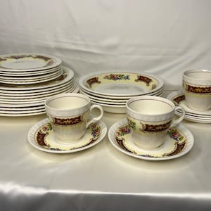 Może przedstawiać: Zestaw vintage porcelany z kwiatowym wzorem, w tym stos talerzy obiadowych, stos talerzy sałatkowych, stos misek do zupy i dwie filiżanki do herbaty z podstawkami.