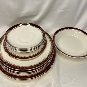 May include: Set of six white ceramic plates with a burgundy and gold rim. The plates are stacked in a pyramid shape.