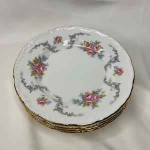 Op de afbeelding: Set van vier witte porseleinen dessertborden met een gouden rand en een bloemmotief met roze rozen en grijze krulwerk.