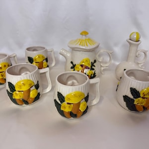 May include: A ceramic tea set comprising a teapot, creamer, sugar bowl, and six mugs. The set is white with a yellow and brown mushroom design. The teapot has a yellow lid, and the mugs have handles.