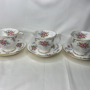 Puede incluir: Tres tazas de té blancas y platillos con un patrón floral. Las tazas y los platillos tienen un diseño delicado y femenino con flores rosas y azules.