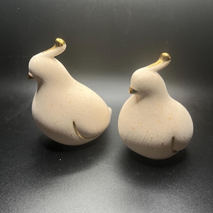 Peut inclure: Deux figurines d'oiseaux en céramique. Les oiseaux sont de couleur beige clair avec des accents dorés sur la tête et le corps. Les figurines ont une forme lisse et arrondie et sont placées sur une surface sombre.