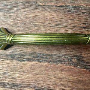 Peut inclure: Une poignée de tiroir décorative de style antique avec une finition en laiton. La poignée présente une poignée centrale cannelée et des extrémités en forme d'éventail. La quincaillerie mesure environ 13 cm de long, adaptée aux meubles.