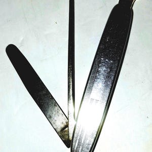 May include: A multi-tool pipe smoking accessory featuring a polished silver-tone metal finish. The tool includes three distinct folding implements: a flat tamper, a thin reamer, and a wider scraping blade, all connected to a central pivot point.