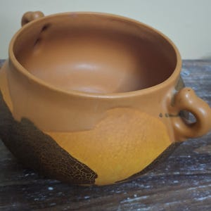 May include: A ceramic bowl with a wide opening and two small handles. The bowl features a color scheme of brown, tan, and yellow, with a textured, crackled pattern on the lower portion. The bowl is likely used for serving food.