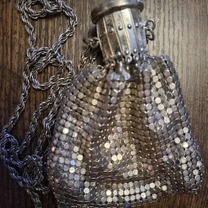 May include: A silver-toned metal mesh pouch featuring a structured, cylindrical top closure with vertical ridges and a matching twisted metal chain strap. The flexible body is composed of small, interlocking circular metal plates.
