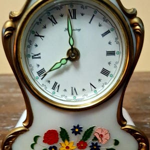 May include: A vintage white porcelain clock with gold trim and floral accents. The clock face has black Roman numerals and green hands. The base features a colorful floral design with red, yellow, and blue flowers. The clock is approximately 15 cm tall.