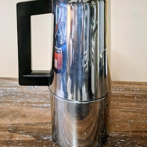 May include: A stainless steel stovetop coffee maker with a polished, reflective finish. The cylindrical body features a black plastic handle attached to the side and a black knob on the lid. The design includes a tapered upper section and a straight base.