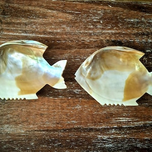 May include: Two fish-shaped dishes crafted from iridescent mother-of-pearl. The dishes display a natural, pearlescent sheen with white, cream, and gold hues. The fish feature a jagged edge along the base and a tail fin.