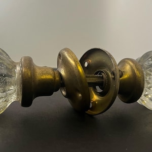 May include: A pair of antique door knobs. The knobs are clear glass with a ribbed design. The base and connecting hardware are brass-coloured metal. The door knobs are for a standard door.