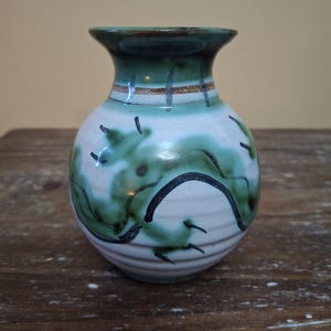 May include: A small, ceramic vase with a rounded body and a flared rim. The vase is primarily white with green and black abstract designs. The rim is dark green, and a thin brown band encircles the neck. The vase sits on a wooden surface.