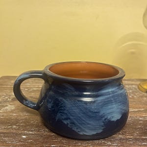 May include: A ceramic mug with a dark blue exterior and a brown interior. The mug has a rounded shape with a handle and a textured, swirled design in shades of blue. The mug is on a wooden surface.