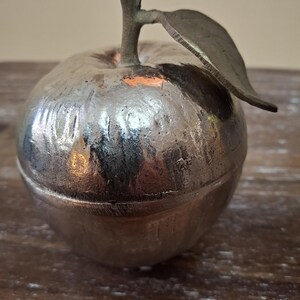 May include: A silver-colored apple-shaped trinket box with a stem and leaf. The box has a textured surface and sits on a wooden surface. The stem and leaf are a darker shade of silver.