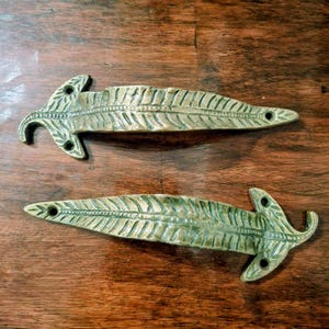 May include: Two elongated, leaf-shaped metal drawer pulls with a textured, antique brass finish. Each handle features a central vein detail, intricate etched foliage patterns, and two circular mounting holes at the base for secure attachment.