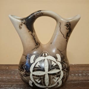 May include: A double-spouted ceramic vessel with a tan and black horsehair finish. The body features a central white etched turtle design with geometric patterns. The two spouts are connected by a bridge handle, creating a distinct silhouette.