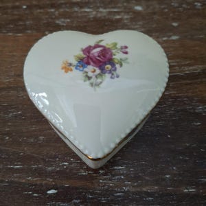 May include: A heart-shaped trinket box. The lid is decorated with a floral design featuring a purple rose and other colourful flowers. The box is white with a gold trim and a beaded edge.