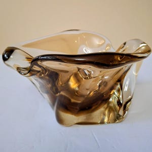 May include: A decorative glass bowl with an unusual, organic form. The bowl has a mix of amber and brown tones, with a shiny finish. The edges of the bowl are softly curved, creating a flowing, artistic design. The bowl is suitable for decorative purposes.