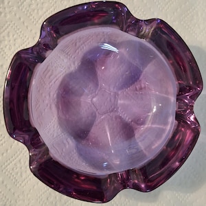 May include: A vintage, purple glass ashtray with a ruffled edge. The ashtray has a textured interior with a central, raised design. The rim is a darker shade of purple than the interior.