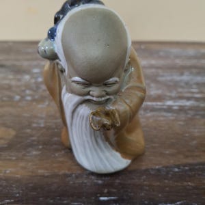 Peut inclure: Figurine en céramique représentant un vieil homme avec une longue barbe blanche et un crâne chauve. La figurine porte une robe marron et porte un petit sac. L'expression du visage est sereine.