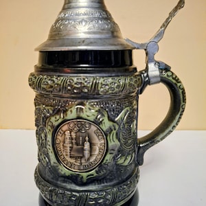 May include: A dark blue ceramic beer stein with a metallic lid and handle. The body features a textured green relief border surrounding a central circular medallion depicting the Munich 1972 Olympic Games with the text XX OLYMPIADE MUNCHEN 1972.