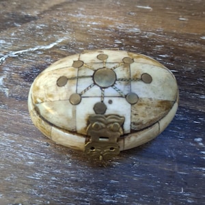 May include: Oval-shaped, antique box with a hinged lid. The box is made of ivory-colored material with a geometric design of brass and metal accents. The clasp is a decorative brass piece. The box is on a wooden surface.