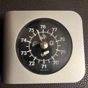 May include: A silver-colored barometer with a black face displaying temperature readings from 70 to 79. The barometer has a round dial with a needle and weather symbols. The device is made in Germany.