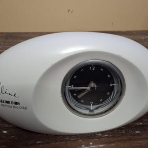 May include: White oval clock with a black clock face. The clock has the name "Celine" and the text "CELINE DION A NEW DAY HAS COME" printed on the front. The clock is on a wooden surface.
