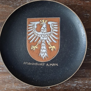 May include: A black ceramic plate featuring the coat of arms of Frankfurt am Main. The emblem includes a white eagle with outstretched wings on a brown shield. The text "FRANKFURT A.MAIN" is below the shield.