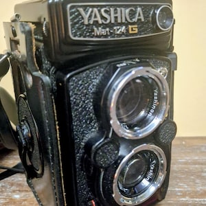May include: A black Yashica Mat-124 G twin-lens reflex camera with a textured body, chrome lens rings, and a black leather case. The front features the Yashica Mat-124 G logo and two Yashinon lenses. The camera has a top-mounted viewfinder hood.