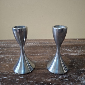 May include: Two silver-colored candle holders with a unique hourglass shape. The base flares out, narrows in the middle, and widens again at the top. The candle holders are made of a shiny metal and sit on a wooden surface.