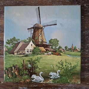 Peut inclure: Carreau de céramique carré représentant un paysage rural avec un moulin à vent, des maisons et une église. Deux cygnes blancs nagent dans un ruisseau. L'œuvre est sur fond de ciel bleu et de paysage verdoyant.