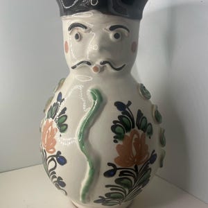 Peut inclure: Pichet en céramique avec un visage, coiffé d'un chapeau noir et d'une moustache peinte. Le corps blanc est orné de motifs floraux orange, verts et bleus, et d'un motif vert en forme de serpent. Le pichet a un aspect artistique unique.