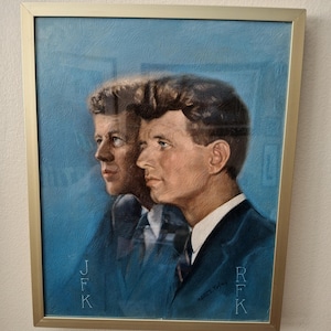 May include: A framed portrait featuring two men in profile against a textured blue background. The left figure is labeled JFK and the right figure is labeled RFK. Both wear dark suits with white collars and ties. The artwork is enclosed in a thin gold frame.
