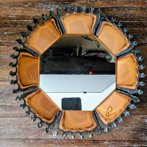 May include: A circular wall mirror featuring an octagonal central glass pane surrounded by a decorative frame with eight petal-like sections in a warm tan color, finished with a dark, jagged, crown-like metal edge and a small signature mark.