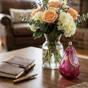 May include: A clear glass vase holds a bouquet of peach roses, white hydrangeas, and lavender sprigs. A pink pear-shaped glass ornament with a leaf detail sits on a wooden table next to a leather-bound journal and a brass pen.