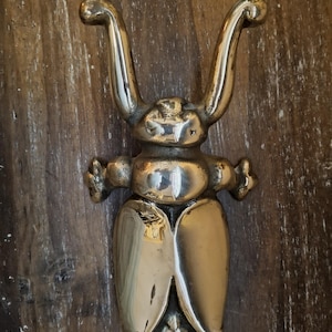May include: A polished brass beetle figurine featuring two long, curved antennae extending upward, a rounded central body, and two large, smooth wing covers. The insect-shaped object has small, decorative protrusions on each side near the head.