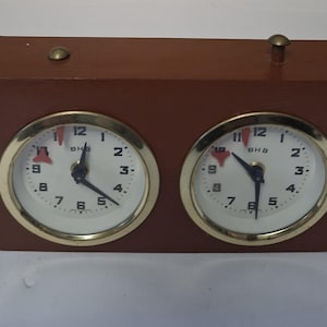 May include: A brown wooden chess clock featuring two circular clock faces with gold-coloured bezels. Each clock face has a white background, black hands, and displays the numbers 1 to 12. The clock is designed for chess or other timed games.