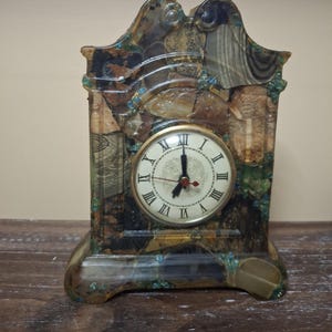 May include: A decorative mantel clock with a cream-coloured face and Roman numerals. The clock's frame is a mosaic of brown, green, and blue tones. The clock hands point to 12:00. The clock sits on a wooden surface.