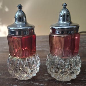May include: A pair of vintage salt and pepper shakers. Each shaker features a clear, faceted base, a red-tinted glass body, and a silver-coloured metal lid. The shakers are on a light brown surface.