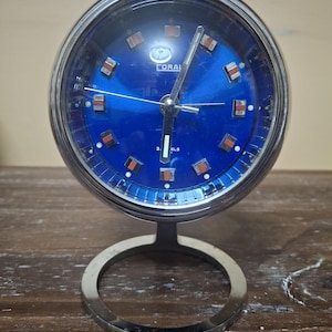 May include: A vintage Coral clock with a round, blue face and silver-tone metal frame. The clock features rectangular hour markers and a silver base. The clock's face has the word "CORAL" printed on it.